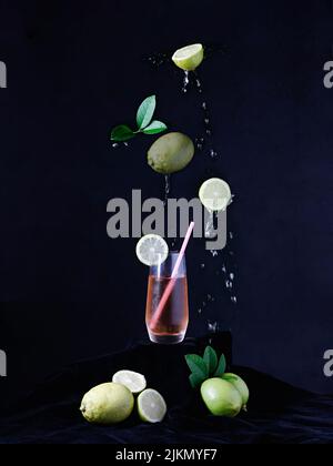 Un colpo verticale di un bicchiere con limoni che cadono su una superficie scura su sfondo nero Foto Stock