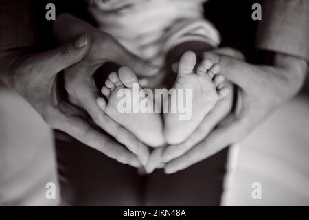 Una bella scala di grigi delle mani di un genitore in una forma del cuore che tiene i piedi del bambino neonato Foto Stock