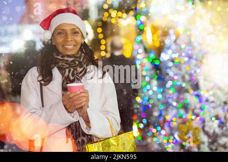 Allegra donna ispanica a Santa Hat con una tazza di tè caldo a piedi alla fiera di strada di Natale Foto Stock