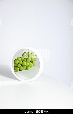 Un bicchierino verticale di uva bianca che si riflette sullo specchio rotondo su sfondo bianco Foto Stock