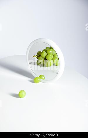 Un bicchierino verticale di uva bianca che si riflette sullo specchio rotondo su sfondo bianco Foto Stock