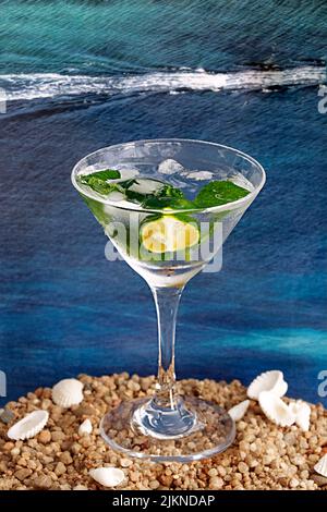 Uno scatto verticale di un bicchiere di cocktail alcolico con calce su una spiaggia di ciottoli con fondo marino Foto Stock