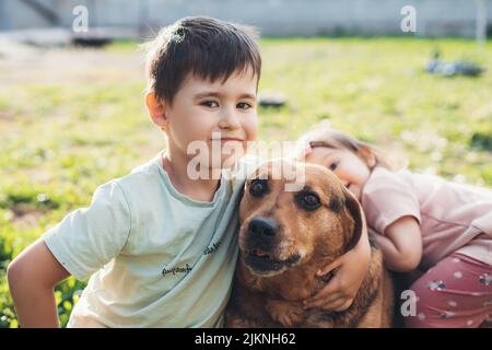 Ragazzo carino e bambina seduta sul prato con il loro cane nel cortile. Ritratto per il design del gioco. Divertimento estivo. Natura estiva. Foto Stock