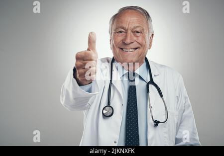 Anni dopo amo ancora il mio lavoro. Un medico anziano in uno studio che dà un pollice su uno sfondo grigio. Foto Stock