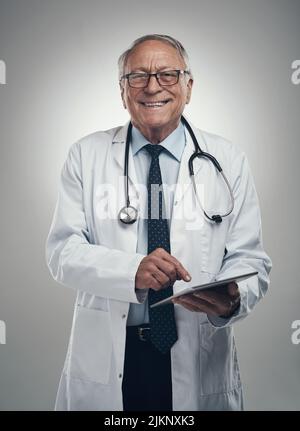 Mi piace vedere un sorriso sui volti dei miei pazienti. Un medico anziano di sesso maschile che usa un tablet digitale in uno studio su sfondo grigio. Foto Stock