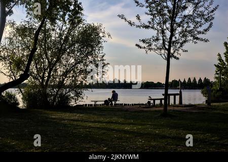 Uomo seduto da solo su una panchina vicino al lago in un parco al tramonto Foto Stock