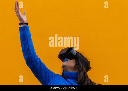 Ritratto isolato di una donna con abiti blu indossando occhiali vr e toccando qualcosa di invisibile in realtà virtuale. Sfondo giallo. Foto Stock