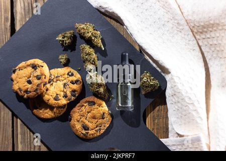 Composizione di biscotti fatti in casa con scaglie di cioccolato, germogli di marijuana essiccati e olio sbd in una bottiglia di vetro con contagocce. Su un bordo di taglio di pietra ambiente Foto Stock