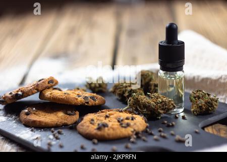 Biscotti fatti in casa con chip di cioccolato con olio cbd, circondati da germogli di cannabis secchi, semi di marijuana e una bottiglia di vetro con un contagocce di olio cbd. Su un Sto Foto Stock