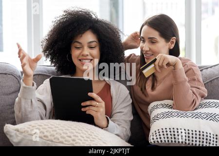 Due giovani donne che acquistano online mentre si siedono sul divano a casa, hanno tanto da comprare, ma sono poco finanziarie. Foto Stock