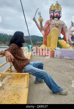 Vista laterale di un attraente giovane uomo con lunghi capelli e barba guardando alla statua del Signore Hanuman mentre si siede sulla panchina accanto al lago in Himachal pradesh Foto Stock