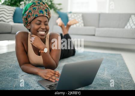 Sorridente giovane ragazza africana am seduta con le gambe incrociate sul tappeto a casa utilizzando il computer portatile e guardando in macchina fotografica Foto Stock