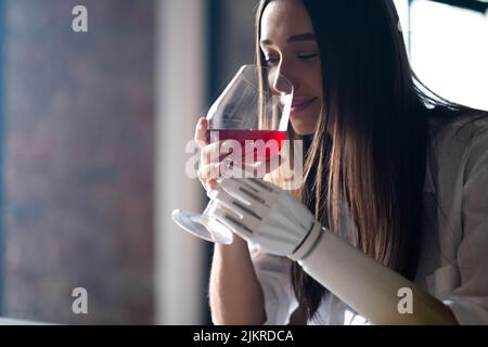 Calmo ragazza premurosa con braccio protesico tiene un bicchiere di vino a casa, donna con disabilità conduce vita normale arto protesico artificiale, persone con Foto Stock