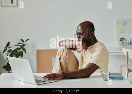 Giovane uomo d'affari africano in occhiali seduti al suo posto di lavoro in ufficio e di lavoro sul laptop Foto Stock