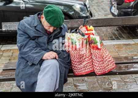 Vecchio uomo seduto su una panca pubblica, annegato sotto la pioggia, tenendo le sue due borse shopping in una mano. Foto Stock