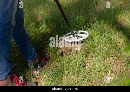 La bobina di un metal detector wireless e le gambe di un uomo in cerca di tesoro su un buco scavato sullo sfondo della foresta. Foto Stock