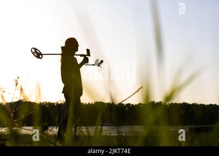 La silhouette di un giovane uomo con un metal detector wireless è un'altra. Sulla riva del fiume, in primo piano l'erba è sfocata Foto Stock