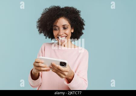 Giovane donna nera sorridente e giocare a gioco online sul cellulare isolato su sfondo blu Foto Stock