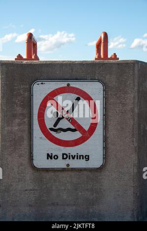 Un primo piano verticale di un cartello in metallo con il testo No Diving su un muro in una spiaggia sotto il cielo luminoso Foto Stock