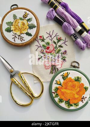 Cerchi, forbici, ago, filo e tessuto vintage in legno da ricamo con cuciture a croce floreali su bianco, vista dall'alto Foto Stock