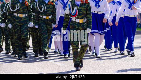 Un gruppo di giovani vestiti di soldati mimetizzazione e uniformi blu e bianche di marinai stanno marciando per le strade della città. Maggio 9. Foto Stock