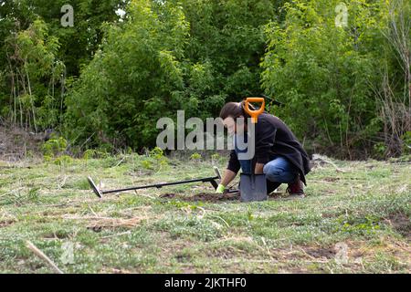 un giovane uomo in jeans, una felpa nera e cuffie wireless sta cercando il terreno in cerca di tesoro utilizzando un pinpointer. nelle vicinanze si trova un wirel Foto Stock