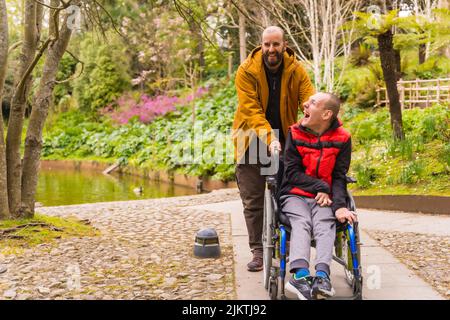 Giovane paralizzato in sedia a rotelle spinto da un amico in un parco pubblico della città Foto Stock