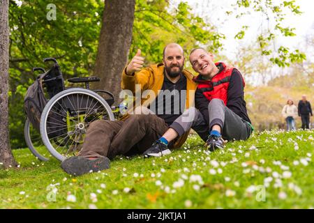 Ritratto di un giovane paralizzato con un amico seduto sull'erba di un parco pubblico in città, parlando e ridendo Foto Stock