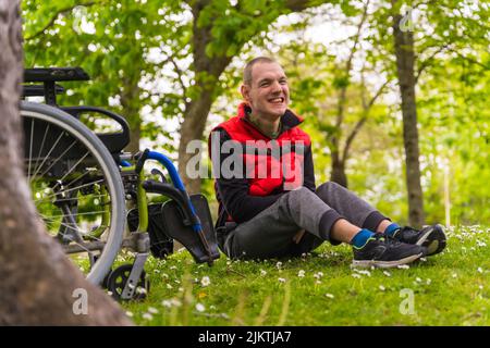 Ritratto di un giovane paralizzato seduto sull'erba accanto alla sedia a rotelle accanto ad un albero Foto Stock