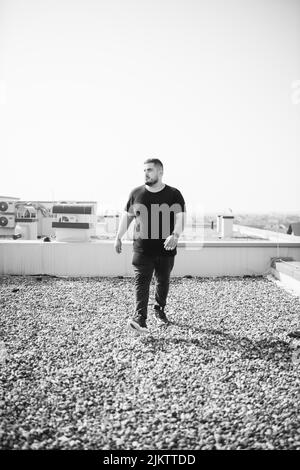 Un colpo in scala di grigi di un cool maschio caucasico in una T-shirt nera che cammina all'aperto con un cielo chiaro sullo sfondo Foto Stock