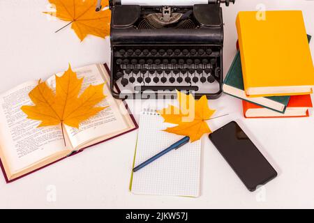 C'è una macchina da scrivere retrò su un tavolo bianco. Nelle vicinanze si trovano libri, notebook con penna, telefono cellulare e foglie di acero giallo. Luogo di lavoro dello scrittore. Foto Stock