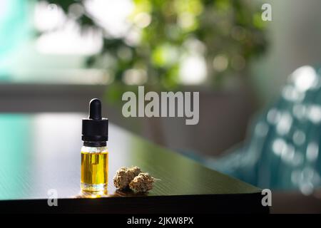 L'olio di marijuana cbd si trova su un tavolo di legno scuro, accanto a due germogli di cannabis secchi. Nello sfondo è un ficus e casa interno in sfocatura. Phot orizzontale Foto Stock