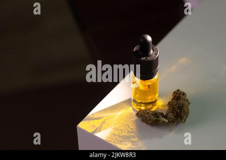 L'olio di marijuana cbd giallo in una bottiglia di vetro con una pipetta si trova su un podio bianco. Nelle vicinanze ci sono due boccioli secchi di cannabis Foto Stock