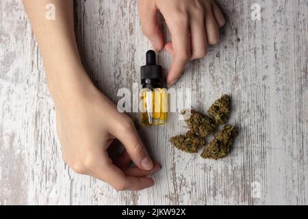 Olio cbd di cannabis in una bottiglia di vetro con una pipetta, boccioli secchi di marijuana medica si trovano nelle vicinanze. Foto con le mani femminili, su sfondo di legno Foto Stock