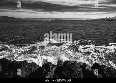 Uno scatto in scala di grigi di un oceano Pacifico ondulato che schiantano le rocce sotto un cielo nuvoloso Foto Stock