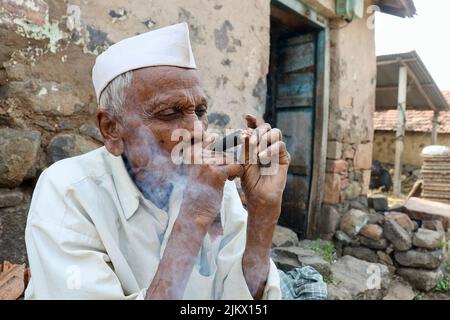 Un fuoco selettivo di un vecchio povero del villaggio indiano in abbigliamento del villaggio che fuma Chillum, anche denominato beedi o bidi o sigaretta fuori della sua casa. Foto Stock