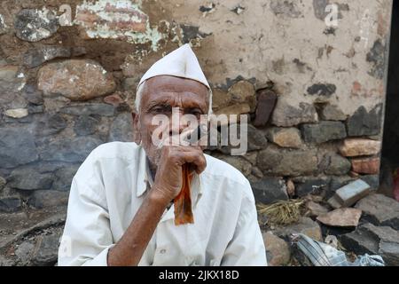 Un fuoco selettivo di un vecchio povero del villaggio indiano in abbigliamento del villaggio che fuma Chillum, anche denominato beedi o bidi o sigaretta fuori della sua casa. Foto Stock