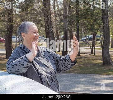 donna anziana con smartphone che parla in conferenza con un amico all'aperto nel parco. giorno dei nonni, giorno della popolazione mondiale, diversità, sel Foto Stock
