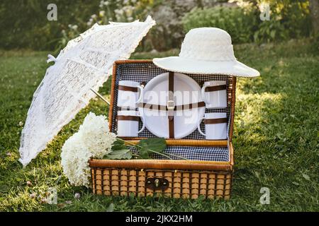 Set romantico con cestino da picnic vintage, ombrellone in pizzo e cappello estivo sul prato del giardino del parco durante le giornate di sole all'aperto. Foto Stock