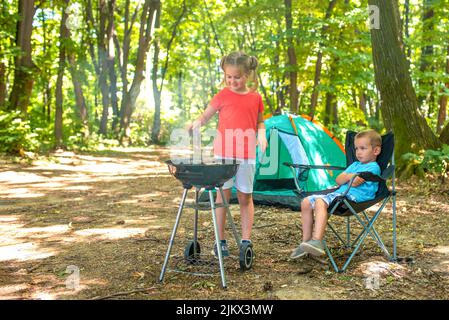 Una sorella caucasica e un fratello che preparano un barbecue in un pic-nic in famiglia nella foresta Foto Stock