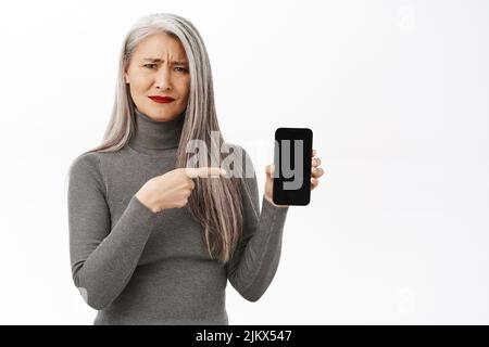 Sconvolto e deluso anziana asiatica, modello coreano di mezza età che punta, mostrando lo schermo dello smartphone e sentendosi triste, sfondo bianco Foto Stock