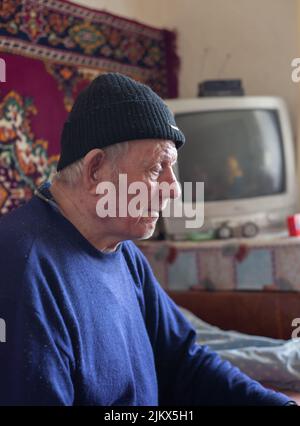 Nonno, un anziano si siede in una casa in un cappello. Sfondo sfocato di vecchi tappeti e TV. Foto Stock