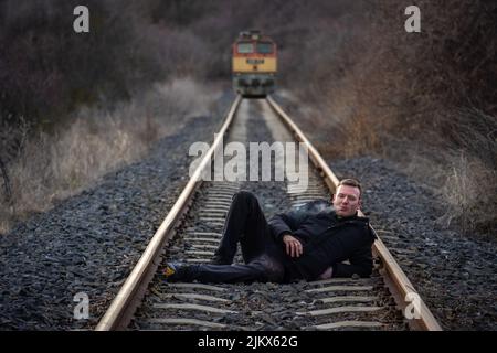 Giovane uomo sdraiato sulla ferrovia che fuma una sigaretta mentre un treno sta arrivando sullo sfondo Foto Stock