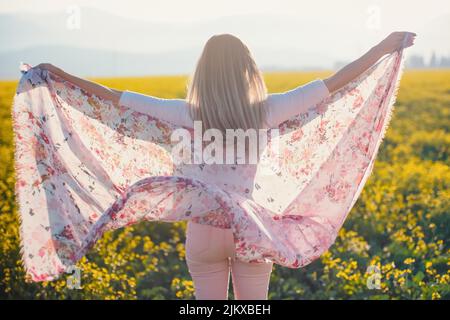 Giovane donna di capelli lunghi ballando in campo di fiori gialli, sciarpa che tiene in movimento dal vento, pomeriggio sfondo luce solare, vista dal retro Foto Stock