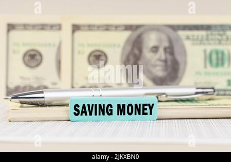 Concetto aziendale. Sui dollari sono una penna e un segno con l'iscrizione - RISPARMIO DI DENARO. Sullo sfondo in uno sfondo sfocato sono dollari Foto Stock