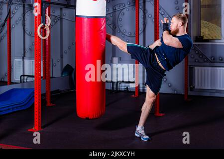 Il giovane lottatore e allenatore di arti marziali miste offre un forte calcio laterale alla borsa da punzonatura rossa. Processo di allenamento in palestra. Foto Stock