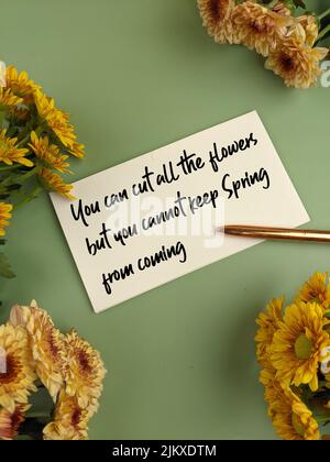 Una vista dall'alto di una nota bianca che dice "si può tagliare tutti i fiori, ma non si può tenere la primavera dal venire", con fiori decorativi e una penna accanto Foto Stock