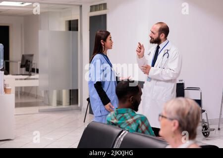 Team medico diversificato che parla di assistenza sanitaria in accoglienza ospedaliera, discutere di pazienti e appuntamenti presso la lobby della struttura medica. Medico e infermiere nella sala d'attesa. Foto Stock