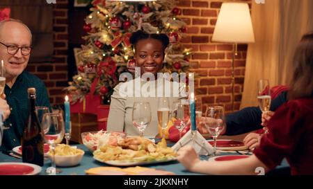 Giovane donna afroamericana sorridente a macchina fotografica mentre si godi la cena di Natale con la famiglia. La persona gioiosa si sente bene mentre festeggia le vacanze invernali con i propri cari. Foto Stock