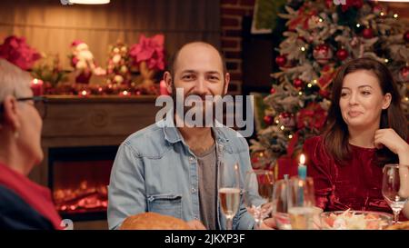 Il marito gioioso sorride alla macchina fotografica mentre si gusta la cena di Natale con i propri cari. Persone felici che si rilassano a casa con amici vicini mentre festeggiano insieme le vacanze invernali. Scatto portatile Foto Stock